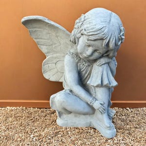 May include: A gray concrete fairy statue. The fairy is depicted in a seated position, with one leg bent and arms wrapped around the other leg. The fairy has large wings and is wearing a dress. The statue is on a bed of small pebbles.