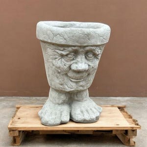 May include: A gray concrete planter shaped like a face with a smiling expression. The planter has two feet and is sitting on a wooden pallet.