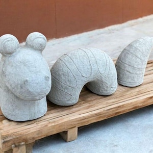 May include: A set of three gray stone sculptures of a frog, a curved shape, and a tail. The sculptures are arranged on a wooden platform.