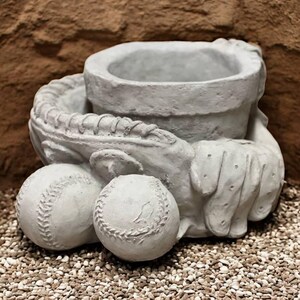 May include: A gray concrete baseball-themed planter. The planter is shaped like a baseball glove holding a baseball and a pot for planting. Two baseballs are at the base of the glove. The planter is on a bed of small pebbles.