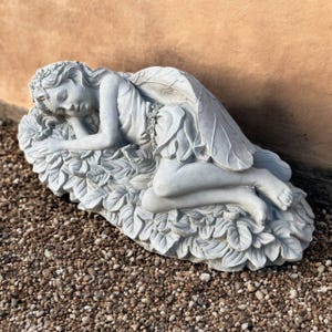 May include: A gray stone fairy statue, sleeping peacefully on a bed of leaves. The fairy has detailed wings and is wearing a leaf-like dress. The statue is a decorative garden ornament.