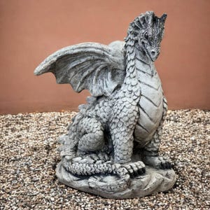 May include: A detailed, gray stone dragon statue with outstretched wings, sitting on a stone base. The dragon has textured scales, a spiked back, and a focused expression. The statue is set against a neutral background.