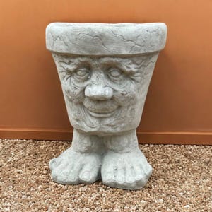 May include: A gray concrete planter shaped like a smiling face with feet. The planter has a textured surface and a wide, smiling mouth. The feet are detailed with toes. The planter is designed for outdoor use.