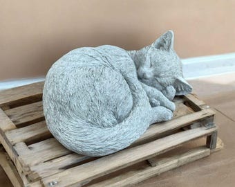 May include: A gray concrete sculpture of a cat curled up sleeping. The cat is lying on a wooden pallet.