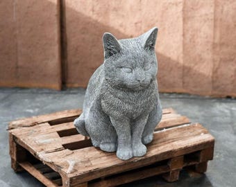 May include: A gray concrete cat statue sitting on a wooden pallet. The cat is sitting with its head slightly tilted and its eyes closed.