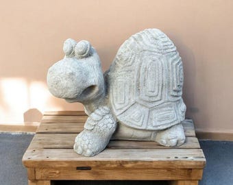 May include: A gray concrete garden statue of a turtle with a whimsical expression. The turtle is standing on a wooden platform.