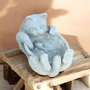 May include: A light gray cat-shaped planter with a fish inside, resting on a rustic wooden stand. The cat is sculpted with its paws forming the sides of the planter. The overall design is whimsical and decorative.