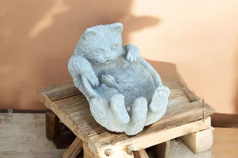 May include: A light gray cat-shaped planter with a fish inside, resting on a rustic wooden stand. The cat is sculpted with its paws forming the sides of the planter. The overall design is whimsical and decorative.