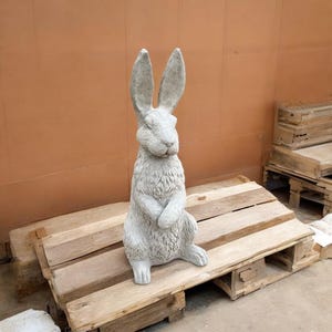 May include: A gray concrete rabbit statue with long ears, sitting upright with its paws crossed. The statue is on a wooden pallet.