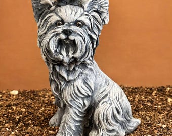 May include: A detailed, gray and white statue of a Yorkshire Terrier dog, sitting upright with a focused gaze. The dog's fur is intricately sculpted, and the statue is set against a brown background.