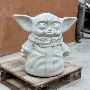 K&ouml;nnte beinhalten: Eine graue Betonstatue eines fiktiven Charakters mit gro&szlig;en Ohren, einer Kapuzenrobe und einem ernsten Ausdruck. Die Statue steht auf einer Holzpalette.