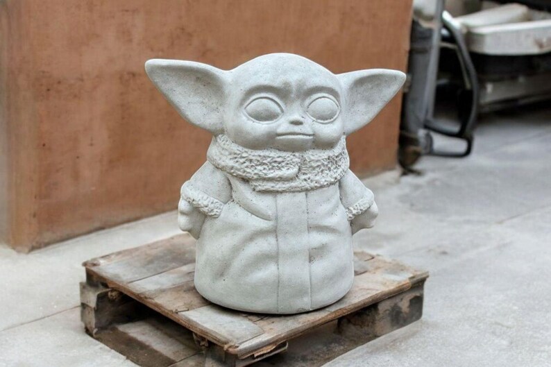 K&ouml;nnte beinhalten: Eine graue Betonstatue eines fiktiven Charakters mit gro&szlig;en Ohren, einer Kapuzenrobe und einem ernsten Ausdruck. Die Statue steht auf einer Holzpalette.