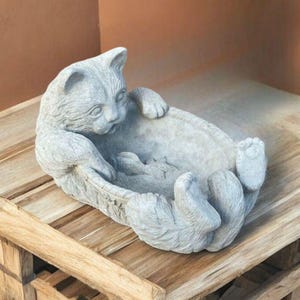 May include: A gray cat-shaped decorative bowl. The cat is lying on its back with its paws in the air, forming the sides of the bowl. The sculpture is made of a stone-like material and sits on a wooden surface.