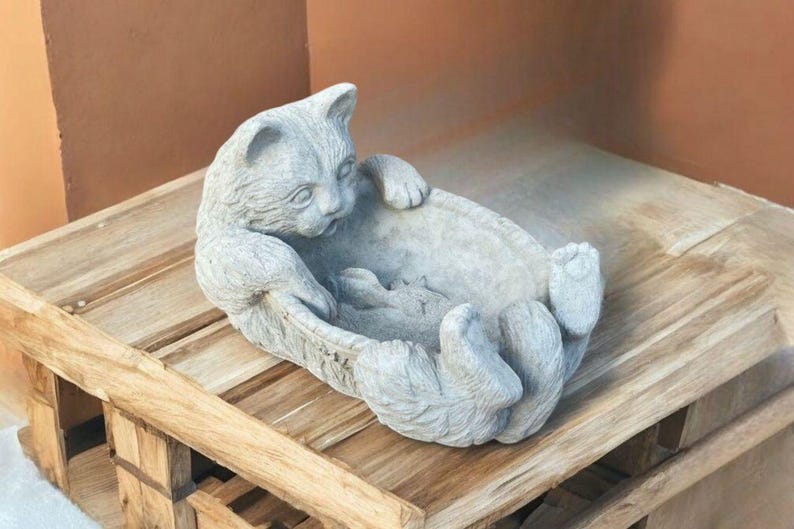 May include: A gray cat-shaped decorative bowl. The cat is lying on its back with its paws in the air, forming the sides of the bowl. The sculpture is made of a stone-like material and sits on a wooden surface.