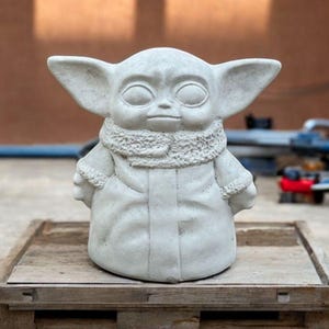 K&ouml;nnte beinhalten: Eine wei&szlig;e Betonstatue eines fiktiven Charakters mit gro&szlig;en Ohren, einem Pelzkragen und einem Mantel. Die Statue steht auf einer Holzplattform.