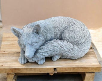Puede incluir: Una escultura de hormigón gris de un zorro acurrucado durmiendo. El zorro está acostado de lado con la cola enroscada alrededor de su cuerpo. La escultura está detallada con textura de pelaje.