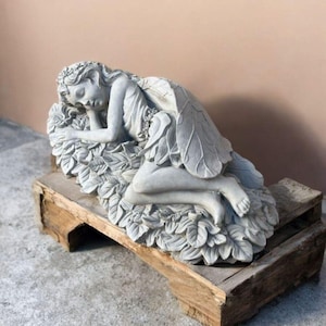May include: A gray stone fairy statue resting on a bed of leaves. The fairy is depicted with closed eyes, wings, and a flowing dress. The statue is placed on a rustic wooden base, creating a serene garden decor piece.