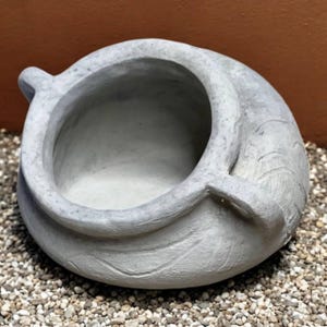 May include: A gray, open-topped, decorative bowl with a textured surface. The bowl has a rounded shape with a curved handle on each side. The bowl is sitting on a bed of small, light-colored pebbles.