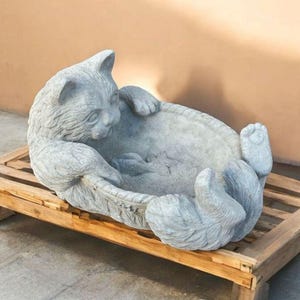 May include: A gray cat-shaped garden statue, lying on its back with paws raised, rests on a wooden pallet. The statue has detailed fur texture and is made of a stone-like material. The cat's face is visible, with its eyes closed.