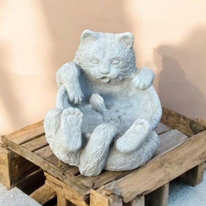 May include: A gray cat statue in a playful pose, seated with its paws in the air. The cat sculpture is made of stone and sits on a rustic wooden crate. The background is a solid, light-colored wall.