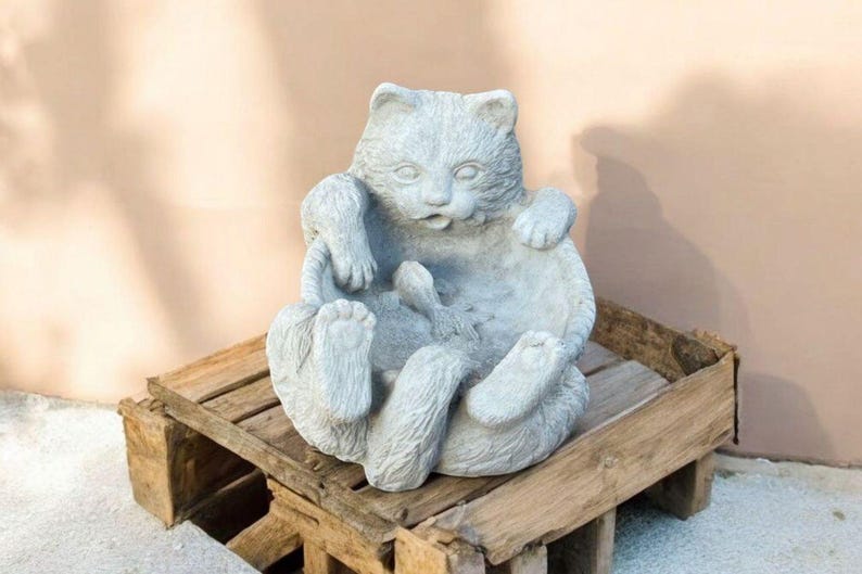 May include: A gray cat statue in a playful pose, seated with its paws in the air. The cat sculpture is made of stone and sits on a rustic wooden crate. The background is a solid, light-colored wall.