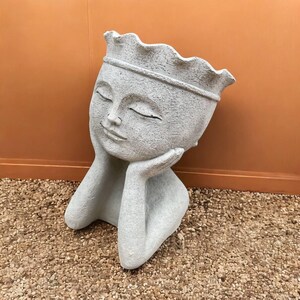 May include: A light gray, sculpted planter shaped like a serene face with closed eyes, resting on its hands. The planter has a crown-like rim and is suitable for small plants or succulents. The overall design is minimalist and elegant.