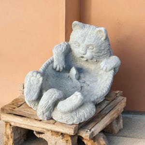 May include: A gray cat sculpture in a relaxed pose, resting in a circular basket-like structure. The cat is sitting on a small, rustic wooden stand. The background is a solid peach-colored wall.