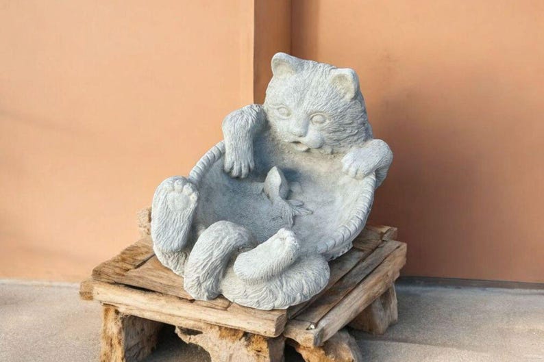 May include: A gray cat sculpture in a relaxed pose, resting in a circular basket-like structure. The cat is sitting on a small, rustic wooden stand. The background is a solid peach-colored wall.
