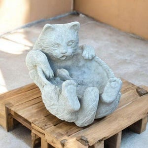 May include: A light gray cat statue in a relaxed pose, lying on its back with paws in the air. The cat sculpture is made of stone and rests on a wooden pallet. The cat's expression is serene.