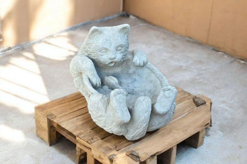 May include: A light gray cat statue in a relaxed pose, lying on its back with paws in the air. The cat sculpture is made of stone and rests on a wooden pallet. The cat's expression is serene.