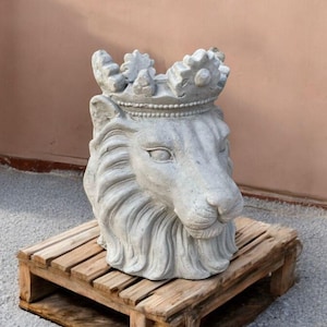 May include: A gray concrete lion head sculpture wearing a crown. The sculpture is placed on a wooden pallet. The lion's head is detailed with fur and facial features. The background is a light brown wall.