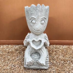 May include: A light gray statue of a character with a smiling face and large eyes, holding a heart shape with its hands. The statue is seated on a log-like base. The background is a solid brown color.