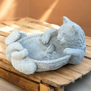 May include: A gray cat statue in a playful pose, resting in a decorative bowl with a fish. The cat sculpture is made of stone and sits on a wooden pallet.