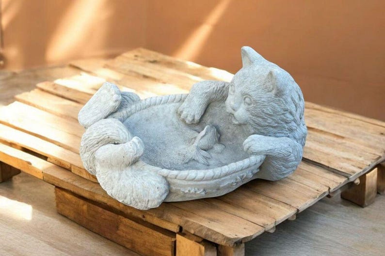 May include: A gray cat statue in a playful pose, resting in a decorative bowl with a fish. The cat sculpture is made of stone and sits on a wooden pallet.