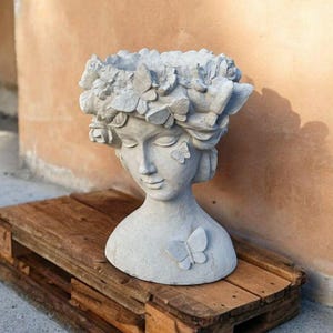 May include: A gray concrete planter shaped like a woman's bust with a floral crown and butterfly accents.