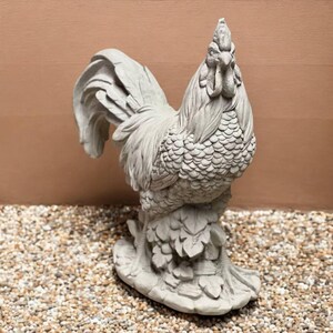 May include: A detailed, gray concrete rooster statue. The sculpture features intricate feather detailing, a prominent comb, and a curved tail. The rooster stands on a base with leaf-like accents, suitable for garden or home decor.