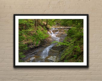 Peut inclure: Photographie encadrée d'une cascade dans une forêt luxuriante. L'eau coule sur des roches sombres, entourée d'un feuillage vert vif. L'image est présentée dans un cadre noir, créant un contraste saisissant avec la scène naturelle.