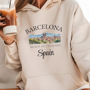May include: Sand-colored hoodie with a front graphic featuring the word "Barcelona" above a watercolor illustration of the city's skyline. Below the image are the words "Region of Catalonia" and "Spain."
