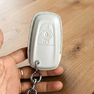 May include: A white leather key fob cover with a silver key ring and clasp. The cover has embossed key symbols and a stitched border. The fob is held against a light wooden surface.
