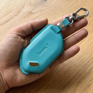 May include: A teal leather key fob case with the embossed text "Queen T". The case has a silver-colored metal clasp and a matching leather strap. The case is held in a hand.