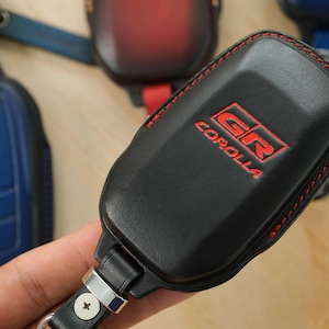 May include: A black leather key fob cover featuring red stitching and the embossed red text GR COROLLA. The cover includes a metal attachment loop with a screw, and other similar key fob covers in blue and red are visible in the background.