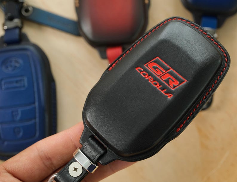 May include: A black leather key fob cover featuring red stitching and the embossed red text GR COROLLA. The cover includes a metal attachment loop with a screw, and other similar key fob covers in blue and red are visible in the background.