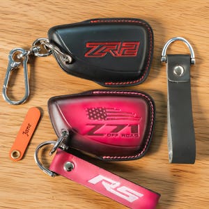 Puede incluir: Una colección de llaveros y llaveros de cuero negro y rojo. Un llavero negro presenta letras rojas "ZR2", mientras que otro llavero rojo muestra "771 OFF ROAD" con un diseño de bandera estadounidense. Una etiqueta naranja está grabada con el nombre "Jane".