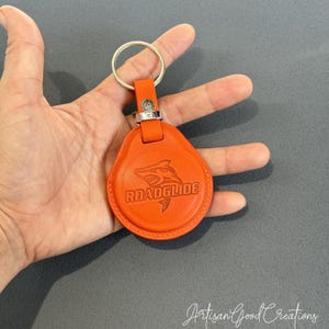 Puede incluir: Llavero de cuero naranja con un llavero plateado. El llavero presenta la palabra "ROADGLIDE" y un gráfico de tiburón. El llavero tiene forma de lágrima y tiene una correa con un cierre de metal plateado.
