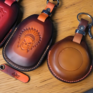 Könnte beinhalten: Leder-Schlüsselanhänger in Braun- und Orangetönen. Einer zeigt ein Totenkopf- und Häuptlings-Design, der andere ein kreisförmiges Design. Jeder hat einen Metallring und einen Lederriemen. Ein kleines Etikett mit dem Namen "Ajay" ist ebenfalls sichtbar.