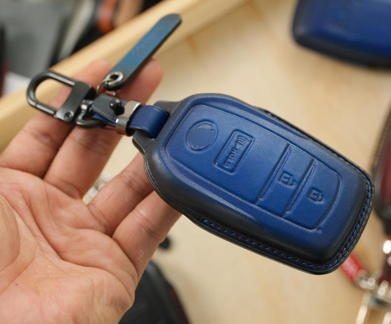 May include: A dark blue leather car key fob cover featuring embossed button icons for locking, unlocking, and holding functions. The cover is attached to a black metal swivel clip and a matching blue leather strap with a silver-toned metal connector.