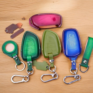 May include: A collection of leather key fobs and covers in various colors including magenta, green, and blue. The items feature metal hardware, including key rings and carabiner clips. One green leather strap is embossed with the name VALERIA.