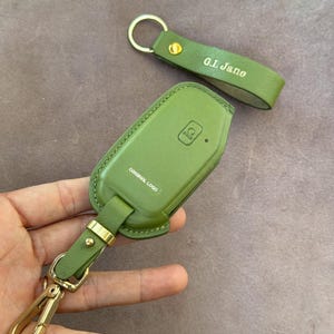 May include: Green leather key fob cover and matching keychain. The key fob cover has the words "ORIGINAL LOGO" printed on it. The keychain has the initials "G.I. Jane" printed on it. Both items have gold-coloured hardware.