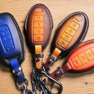 May include: Four oval-shaped leather key fob covers in blue, tan, dark brown, and orange. Each cover features embossed button icons, including a lock, unlock, trunk release, and panic button, with matching leather straps and silver metal hardware.