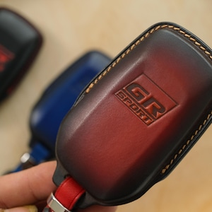 May include: A dark red and black gradient leather key fob cover featuring the embossed text GR SPORT. The cover has visible tan stitching along the edges and is attached to a red leather strap with a silver metal connector at the base.